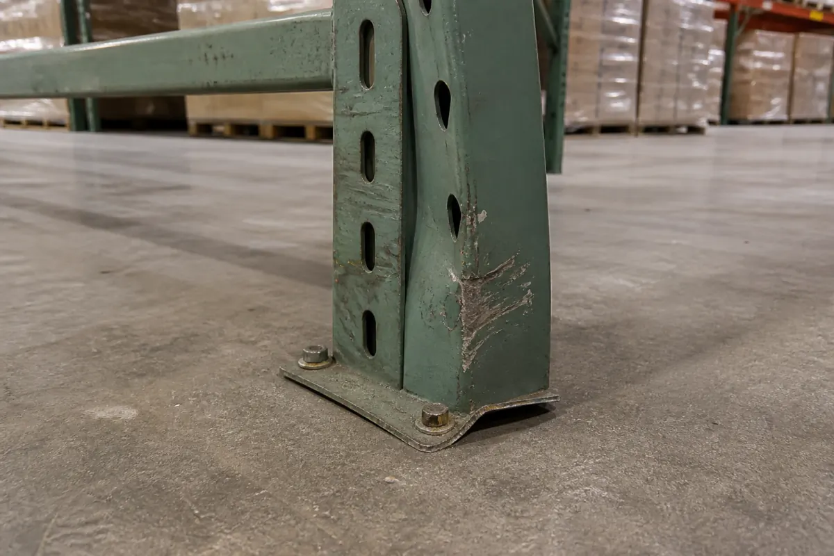 Green pallet rack upright with visible forklift impact damage, a creased and dented column face, and a footplate curling away from the concrete floor