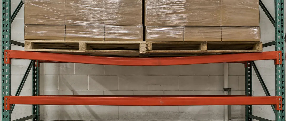 Loaded orange pallet rack beam bowing visibly downward under palletized cartons, compared with a straight unloaded beam on the level below