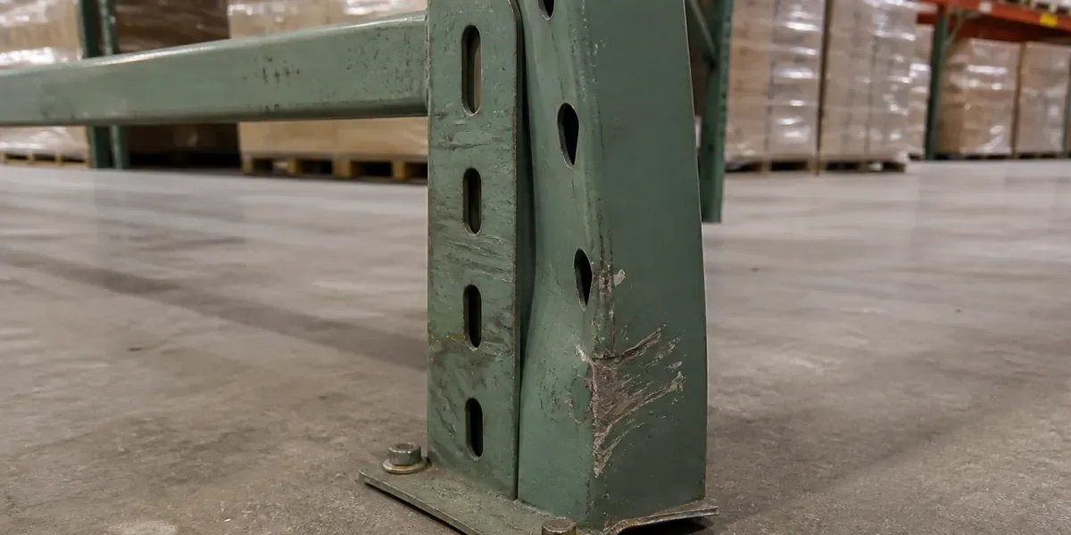 Impact-damaged green pallet rack upright with a dented column and curled footplate on a warehouse floor
