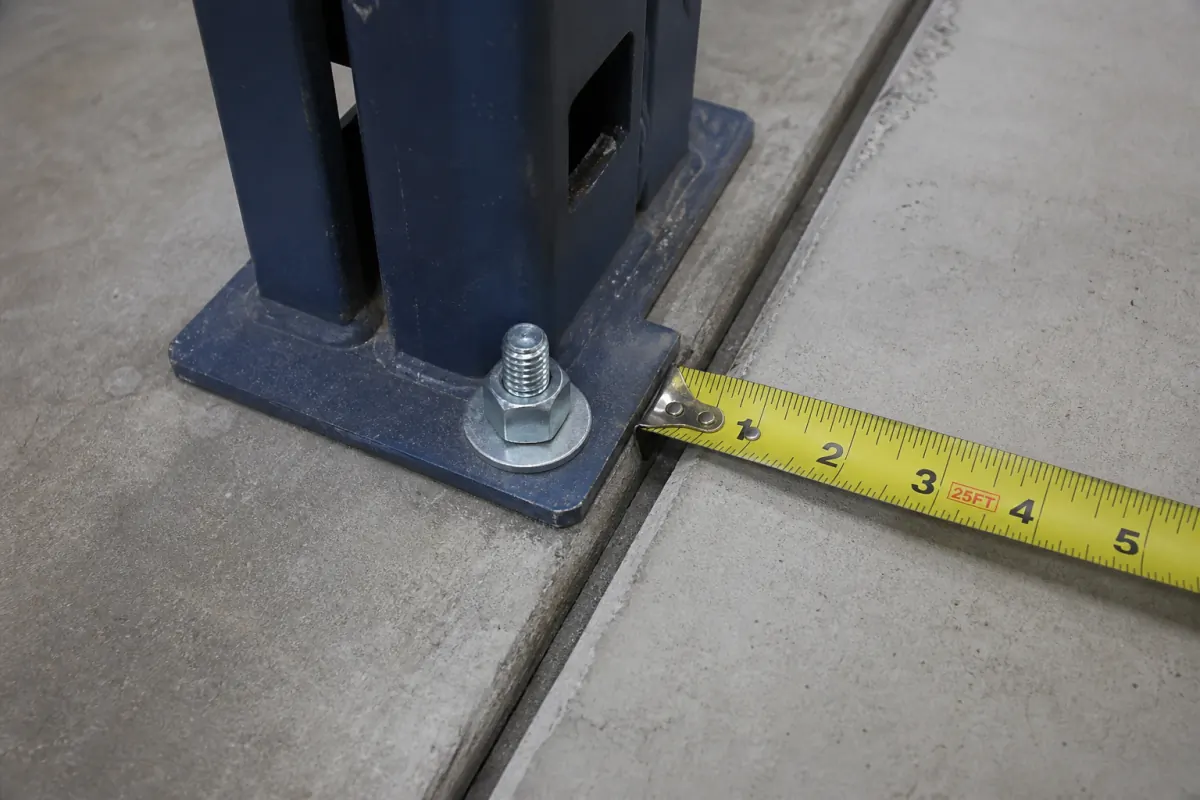 Tape measure showing roughly one inch between a pallet rack anchor bolt and a saw-cut control joint in a concrete warehouse slab
